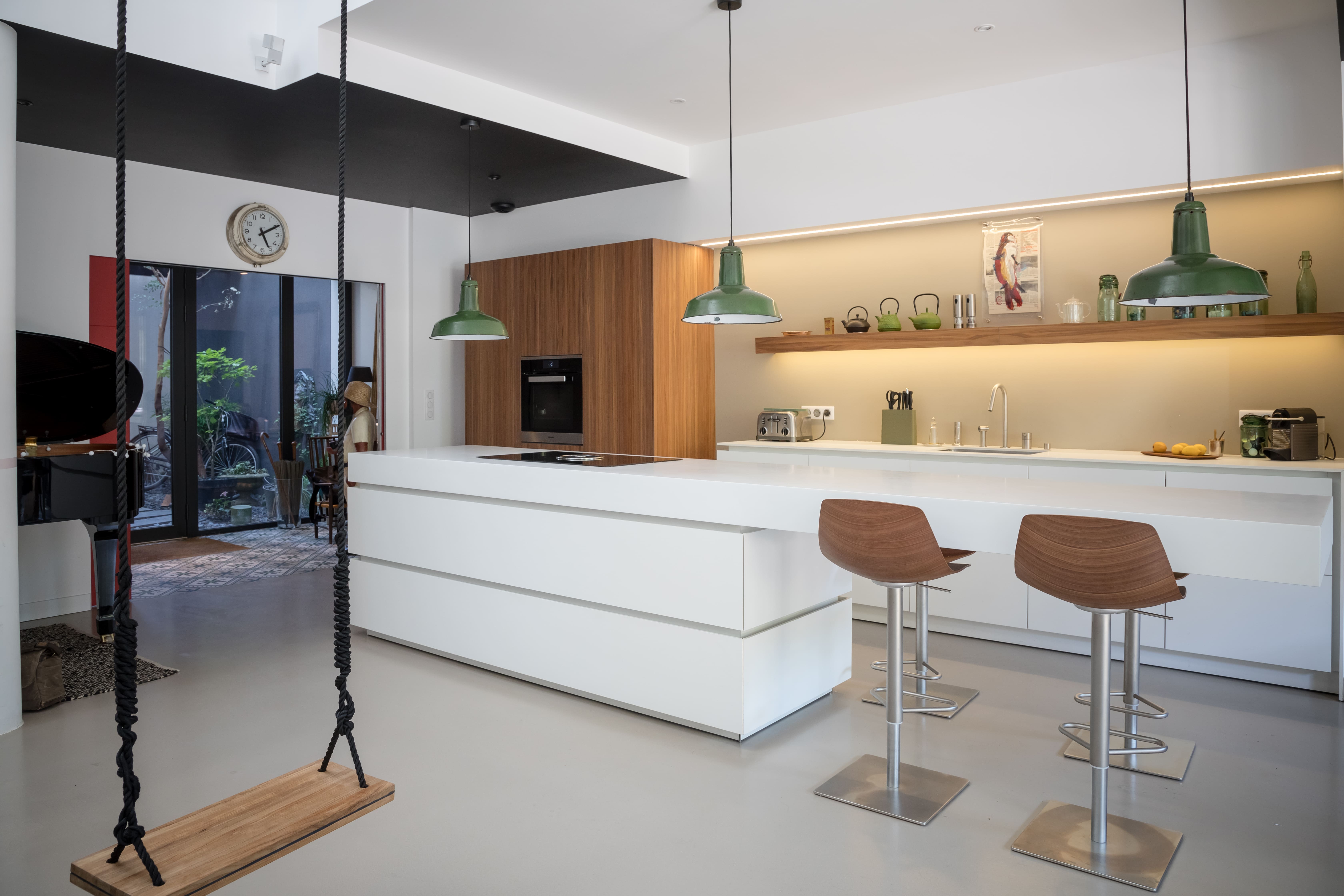 Cuisine design blanche avec étagère en bois, suspensions vertes et bar intégré, mêlant modernité, chaleur et fonctionnalité dans un espace lumineux.