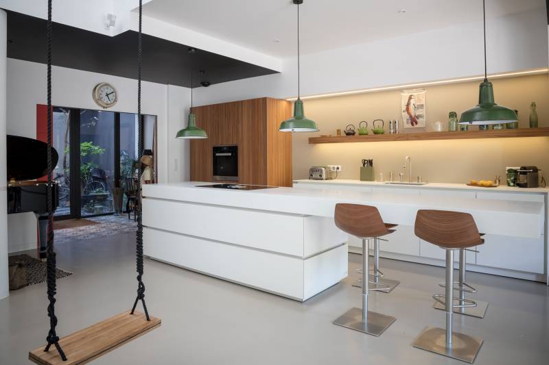 Cuisine design blanche avec étagère en bois, suspensions vertes et bar intégré, mêlant modernité, chaleur et fonctionnalité dans un espace lumineux.
